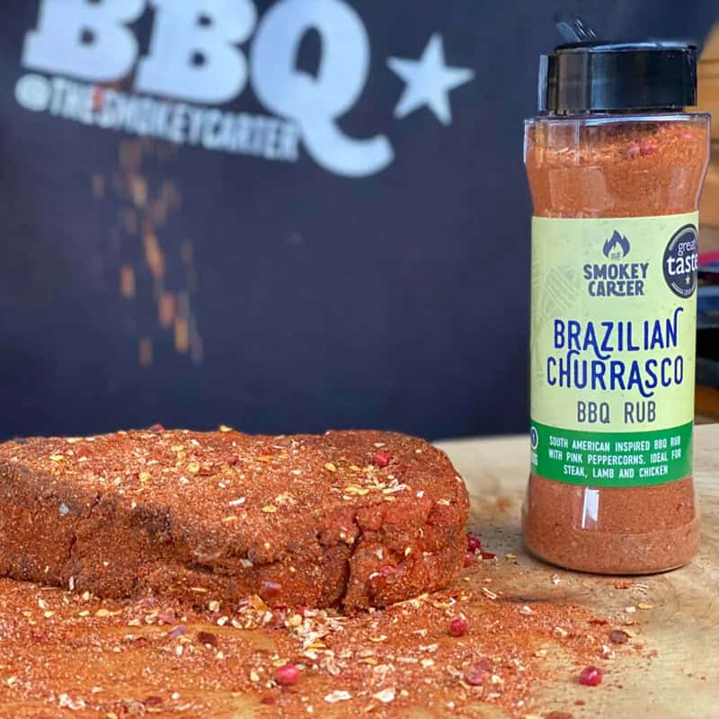Brazilian Churrasco BBQ Rub in a clear spice jar with a black lid, placed next to a seasoned piece of meat on a wooden surface against a dark background.