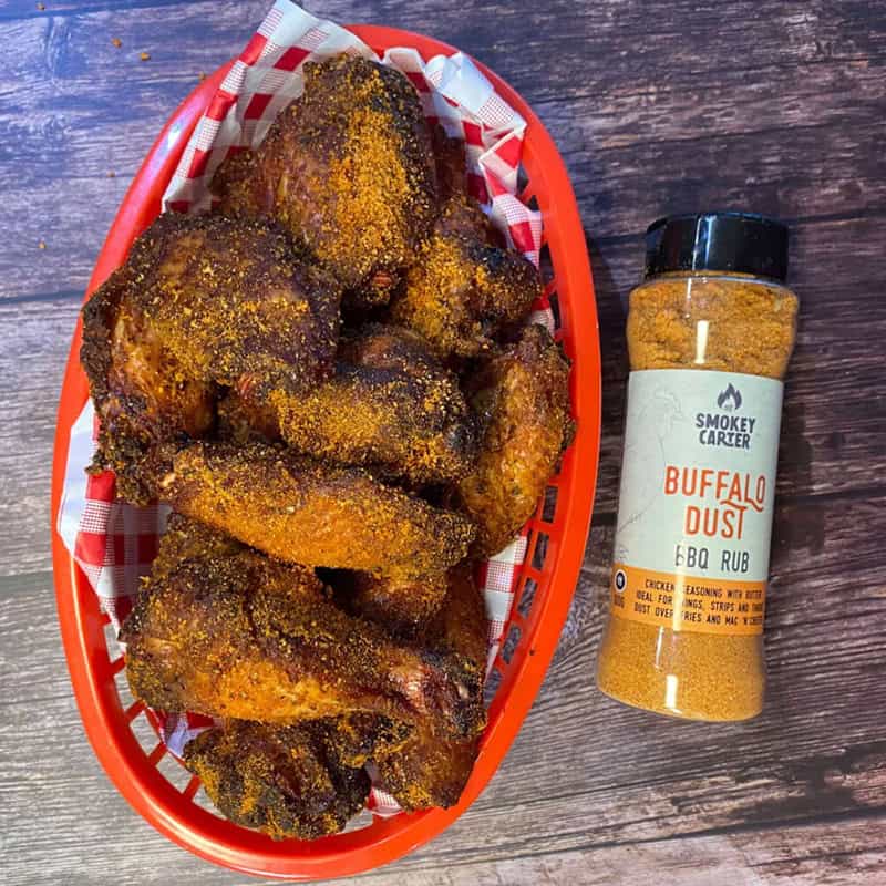 Crispy, seasoned fried chicken wings in a red basket lined with checkered paper next to a bottle of Smokey Carter Buffalo Dust BBQ rub on a rustic wooden table.