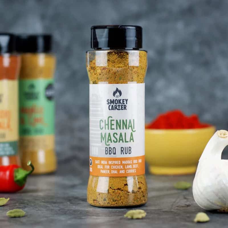 Smoky Carter Chennai Masala BBQ Rub spice jar on a kitchen countertop with spices and herbs, ideal for grilling chicken, lamb, beef, and curries.