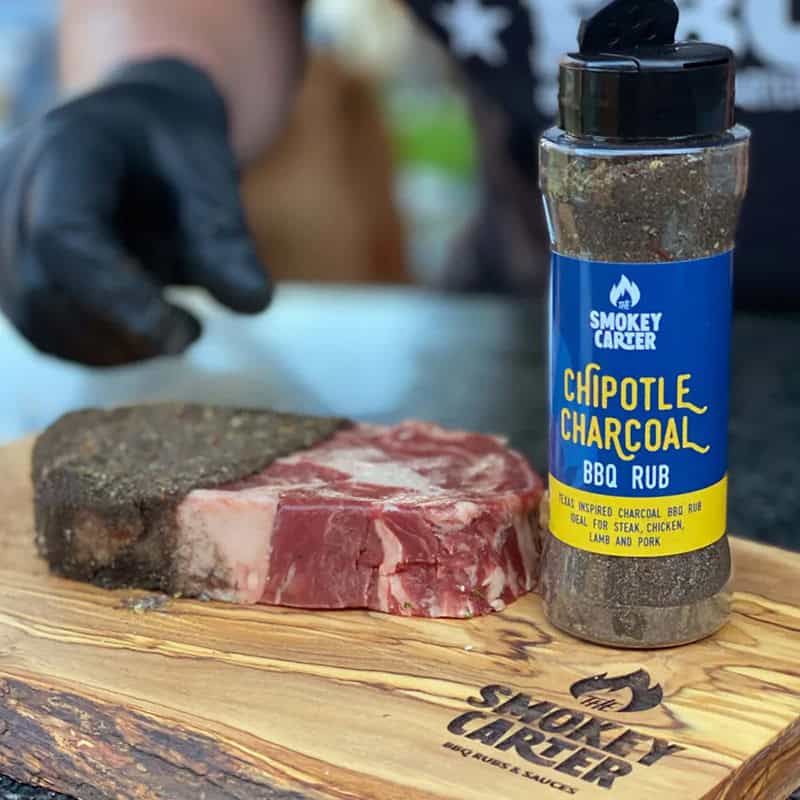 Smokey carter chipotle charcoal BBQ rub on seasoned meat with cedar wood board for grilling or smoking, all-natural wood rub for steak, chicken, lamb, and pork, high-quality charcoal BBQ seasoning.