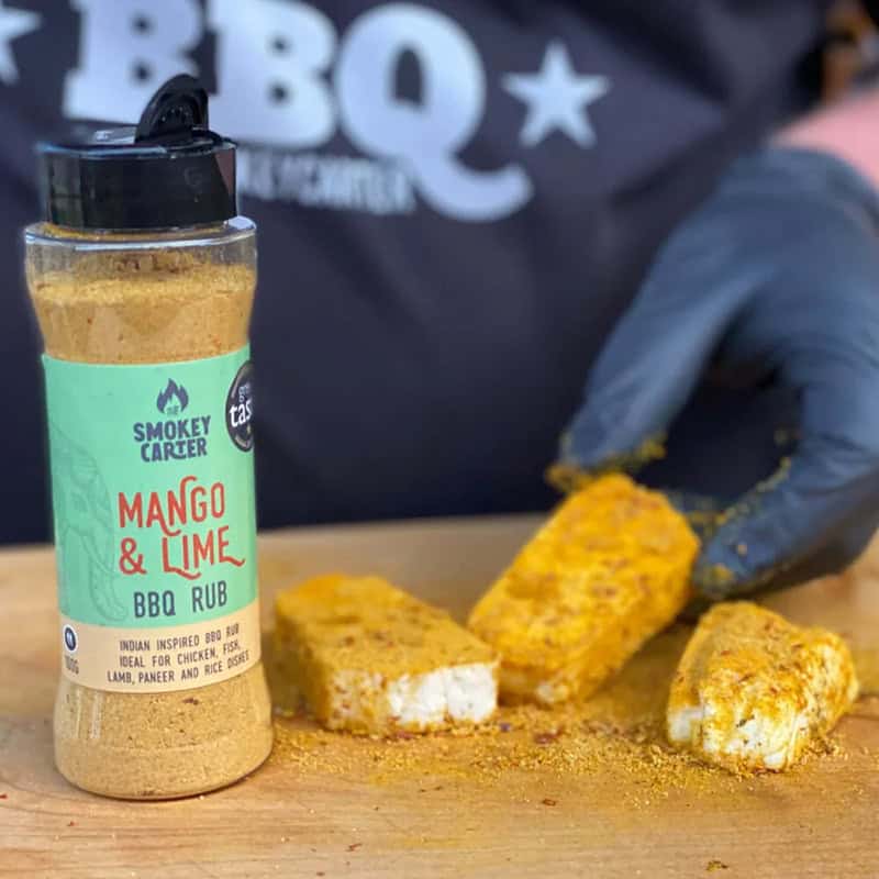 Smokey Carter Mango & Lime BBQ Rub bottle with seasoned chicken bites on a wooden surface, showcasing flavorful grilling spices.