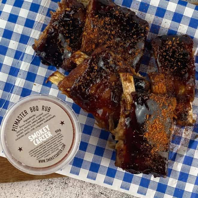 Juicy smoked ribs glazed with BBQ sauce on a blue checkered paper, accompanied by a container of smoky BBQ rub, highlighting Cedar Works' expertise in grilling and meat preparation.