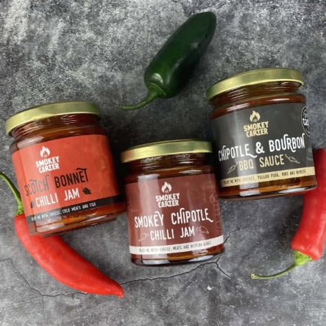 Savory smoked chili jams and BBQ sauce jars on a textured gray surface with fresh chili peppers and a jalapeño, showcasing Cedar Works’ artisanal condiments and gourmet food products.