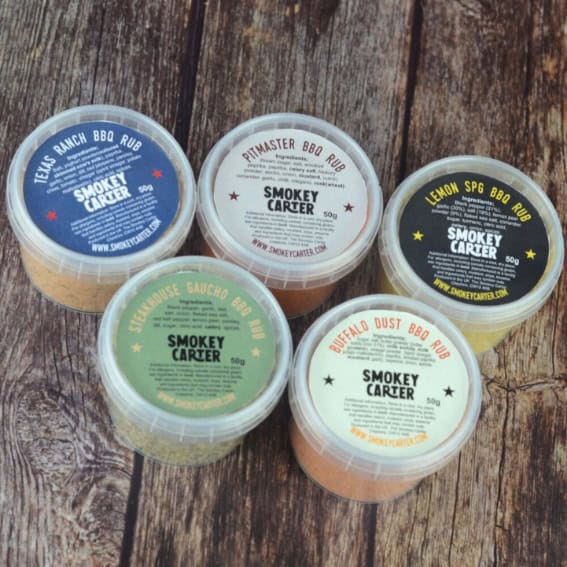 Spicy barbecue rubs in small containers from Smokey Carter, including Texas Ranch, Pitmaster, Lemon Spg, Szechuan Saucha, and Buffalo Dust, displayed on rustic wooden surface.