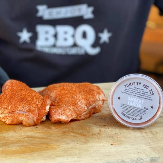 Smoked chicken thighs seasoned with cedarwood spices on a wooden cutting board with Pitmaster BBQ Rub container, showcasing gourmet BBQ preparations.