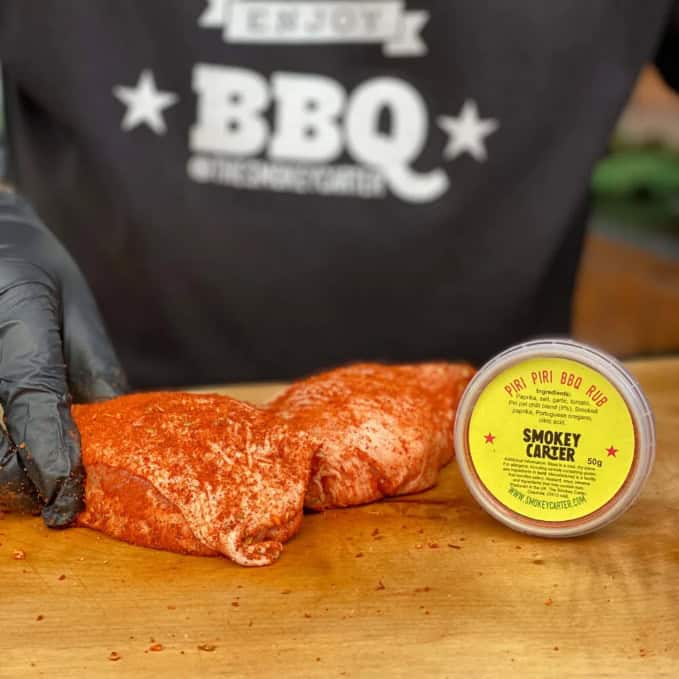 Smokey Carter Piri Piri BBQ Rub with seasoned raw chicken breast on wooden cutting board, Chef preparing BBQ cooking, Cedar Works logo in background, outdoor grilling scene.