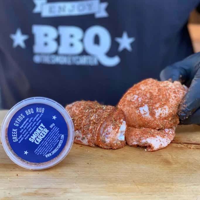 image054 Juicy smoked chicken wrapped with spices on a wooden cutting board, with a container of Greek Gyros BBQ rub sauce nearby, in front of a person wearing a Cedar Works BBQ apron.