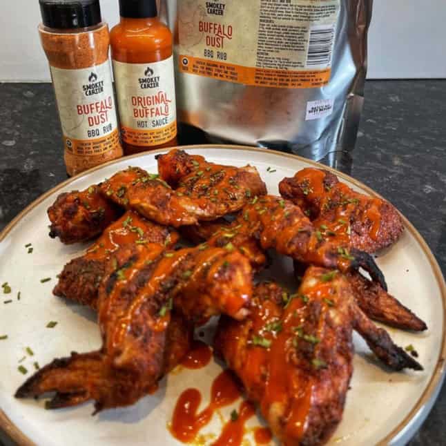 Smokey Carter BUFFALO HOT SAUCE_3 Buffalo wings with spicy hot sauce and fresh herbs served on a white plate, with bottles of smoky barbecue sauce and seasoning in the background. Perfect for grilling, barbecues, and outdoor cookouts.