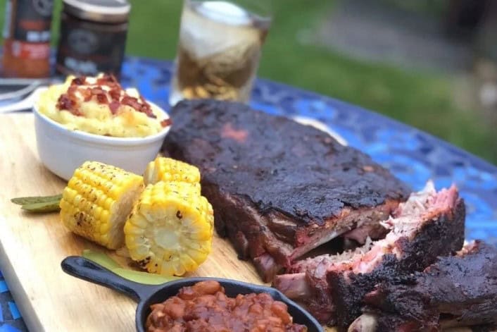 Smokey Carter Chipotle bourbon bbq sauce 5 Smoked barbecue ribs with grilled corn on the cob and side dishes on a cedar wood platter outdoor dining image.