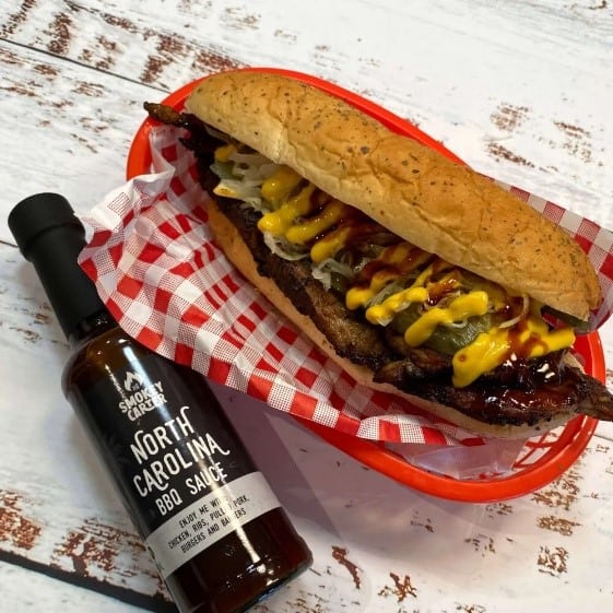 Juicy BBQ brisket sandwich with mustard and pickles in a toasted bun, served in a red basket with checkered paper, accompanied by North Carolina BBQ sauce for enhanced flavor.